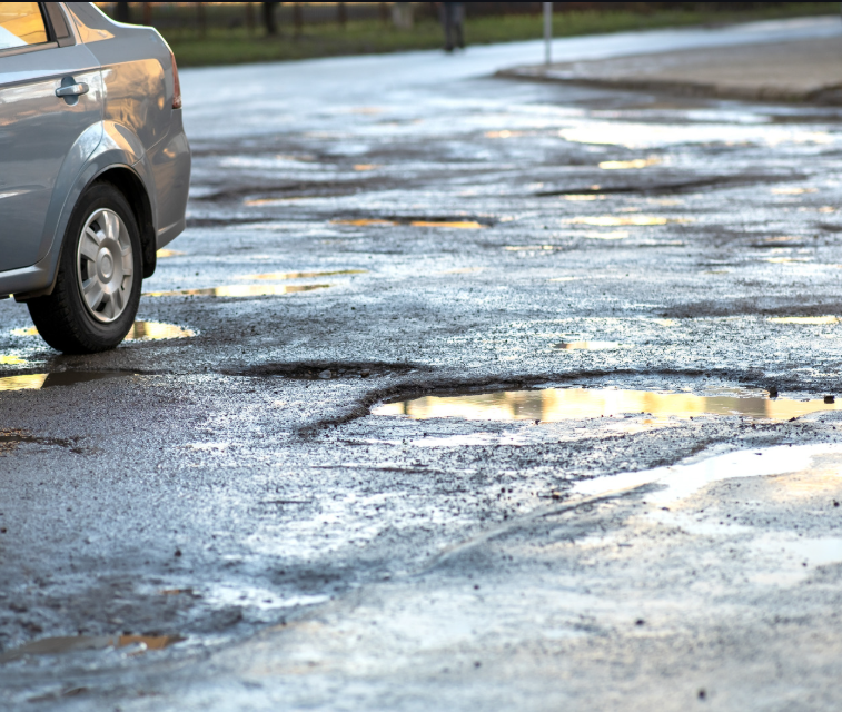 A gray car navigates a pothole-filled, wet road - https://www.dbennettinsurance.com/
